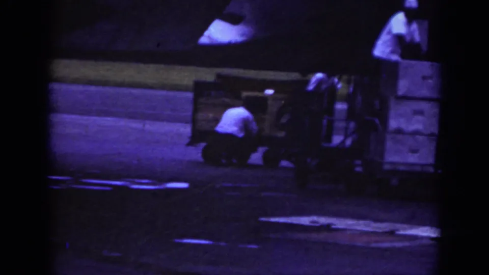 1962 Raleigh tarmac view with ground crew crouched at a baggage trailer near an aircraft edge, filmed in soft 8mm color.