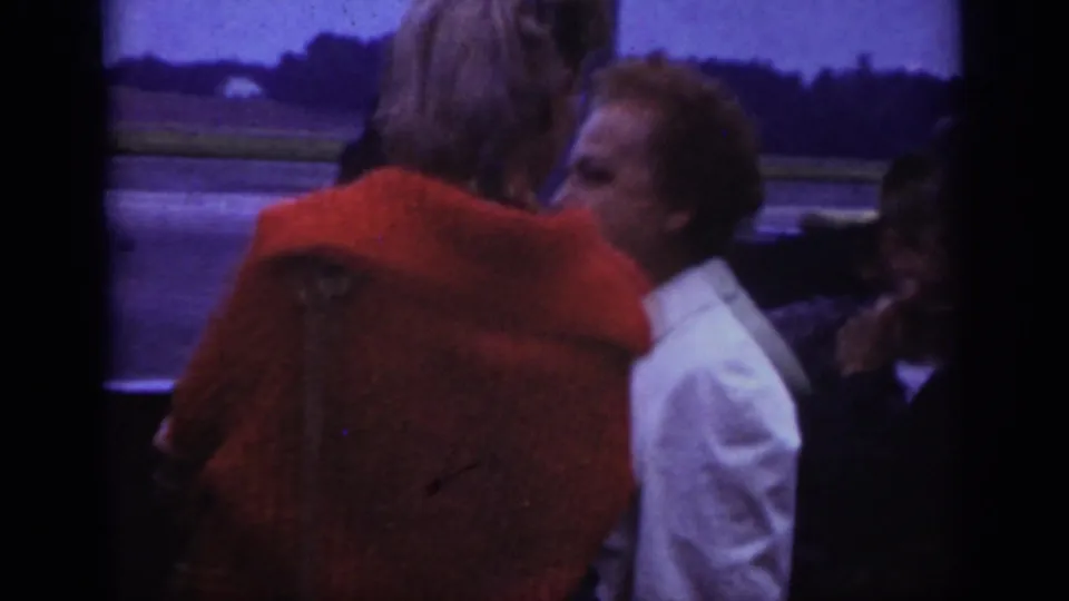1962 Raleigh airport frame showing passengers waiting near the tarmac, with a red coat and light jacket prominent in the foreground.