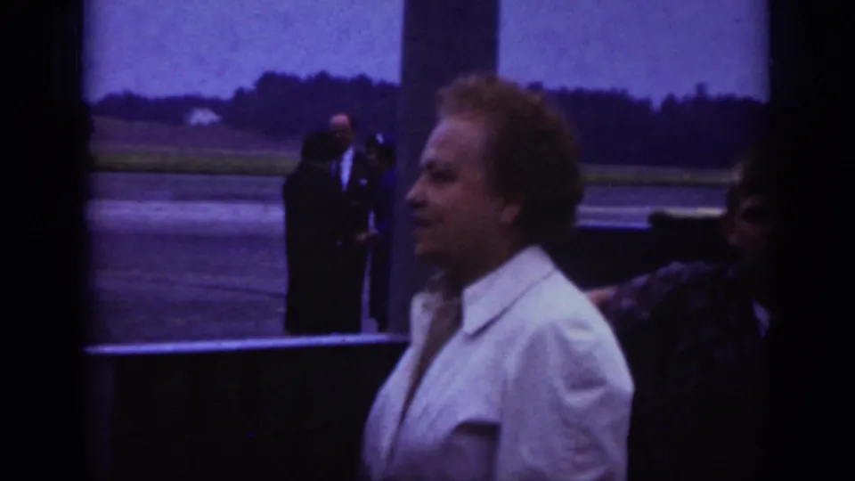 1962 Raleigh airport home-movie frame with a man in a light jacket near other passengers and open runway behind him.