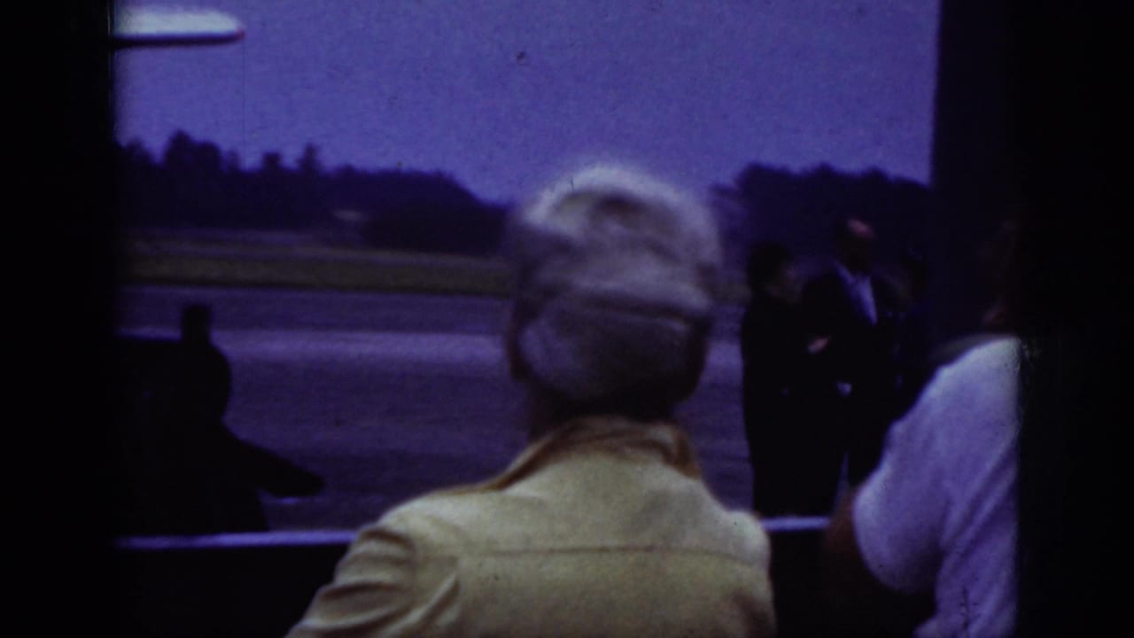 1962 Raleigh airport home-movie frame showing adults watching from a shaded area toward people on the tarmac.
