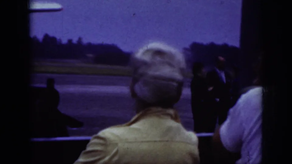 1962 Raleigh airport home-movie frame showing adults watching from a shaded area toward people on the tarmac.