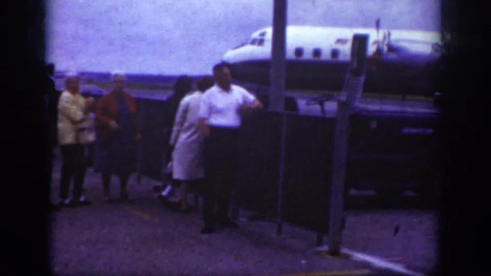 1962 Raleigh airport home-movie frame showing people near a tarmac barrier with an aircraft and boarding stairs behind them.