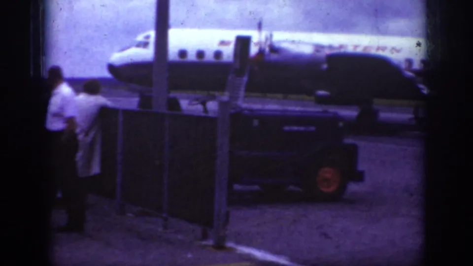 1962 Raleigh airport frame with a dark vehicle crossing the foreground near spectators and an Eastern Airlines aircraft on the tarmac.