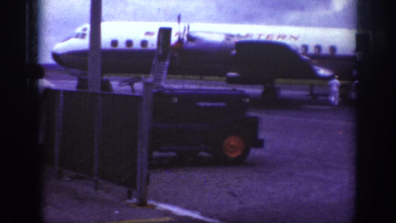 1962 Raleigh airport home-movie frame showing a Western Airlines propeller plane, mobile stairs, ground crew, and utility vehicle on the tarmac.