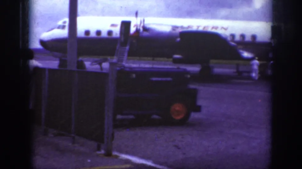 1962 Raleigh airport home-movie frame showing a Western Airlines propeller plane, mobile stairs, ground crew, and utility vehicle on the tarmac.