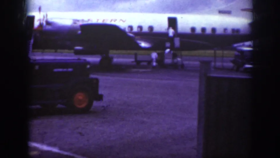 Home-movie frame from Raleigh in 1962 showing the Western Airlines fuselage, service stairs, crew members, and a dark airport vehicle.