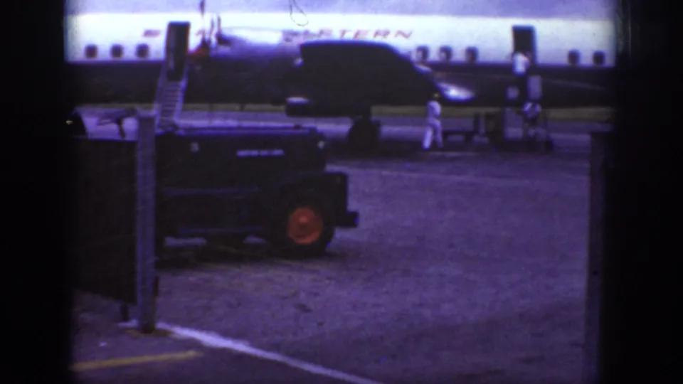 1962 Raleigh airport frame showing a Western Airlines aircraft being attended by ground crew beside boarding stairs and a dark tarmac vehicle.