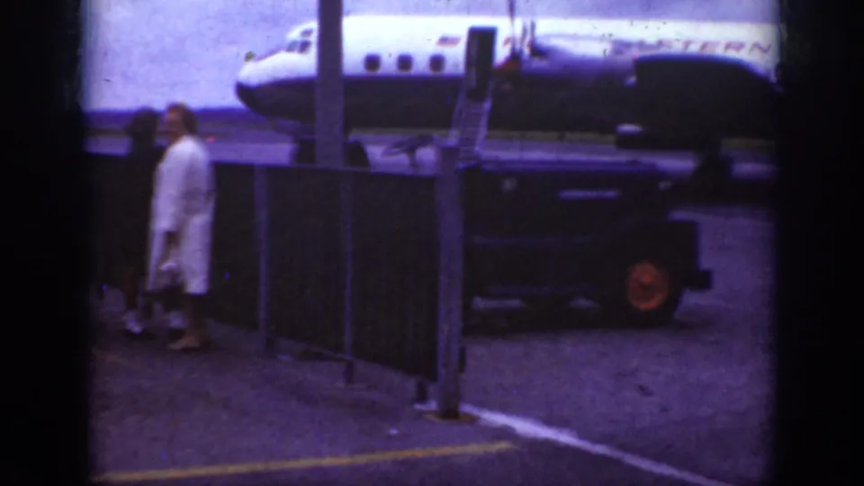 Traveler in a light coat crosses the Raleigh airport ramp near fencing, with the Western Airlines aircraft and boarding stairs behind.