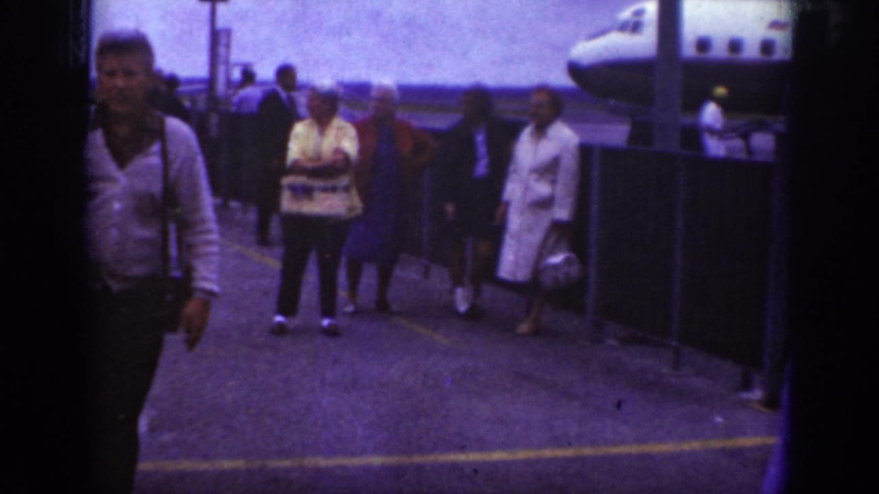 1962 Raleigh airport home-movie frame showing travelers on the tarmac near an aircraft, with several people walking or waiting by a railing.
