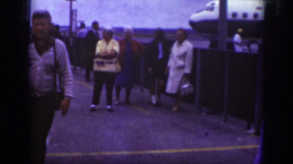 1962 Raleigh airport home-movie frame showing travelers on the tarmac near an aircraft, with several people walking or waiting by a railing.