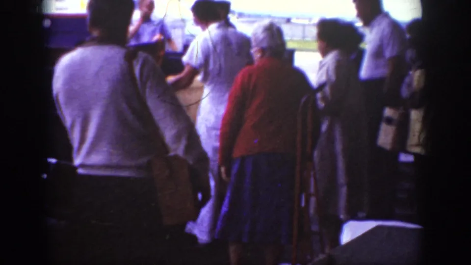 Home-movie frame from 1962 Raleigh showing a small group of travelers moving toward boarding, with period clothing and handheld 8mm softness.
