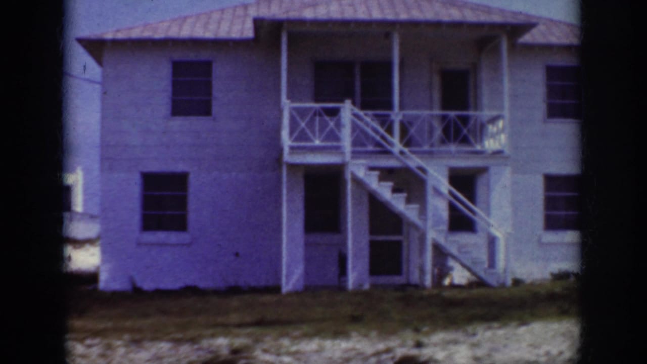 1962 Raleigh home-movie frame showing a white two-story residence with exterior stairs, porch railings, dark windows, and sandy ground in front.