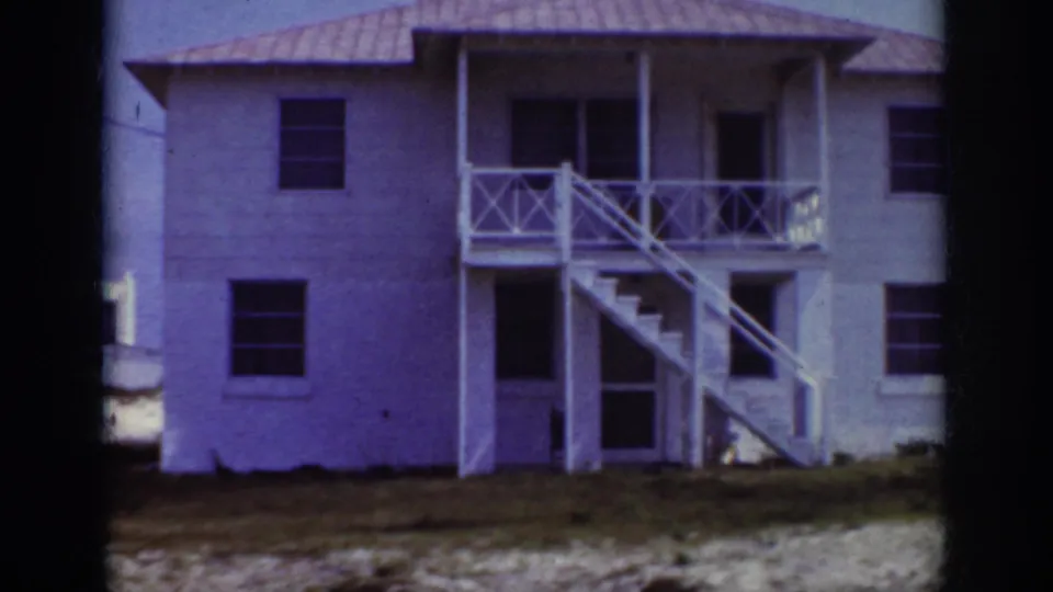 1962 Raleigh home-movie frame showing a white two-story residence with exterior stairs, porch railings, dark windows, and sandy ground in front.