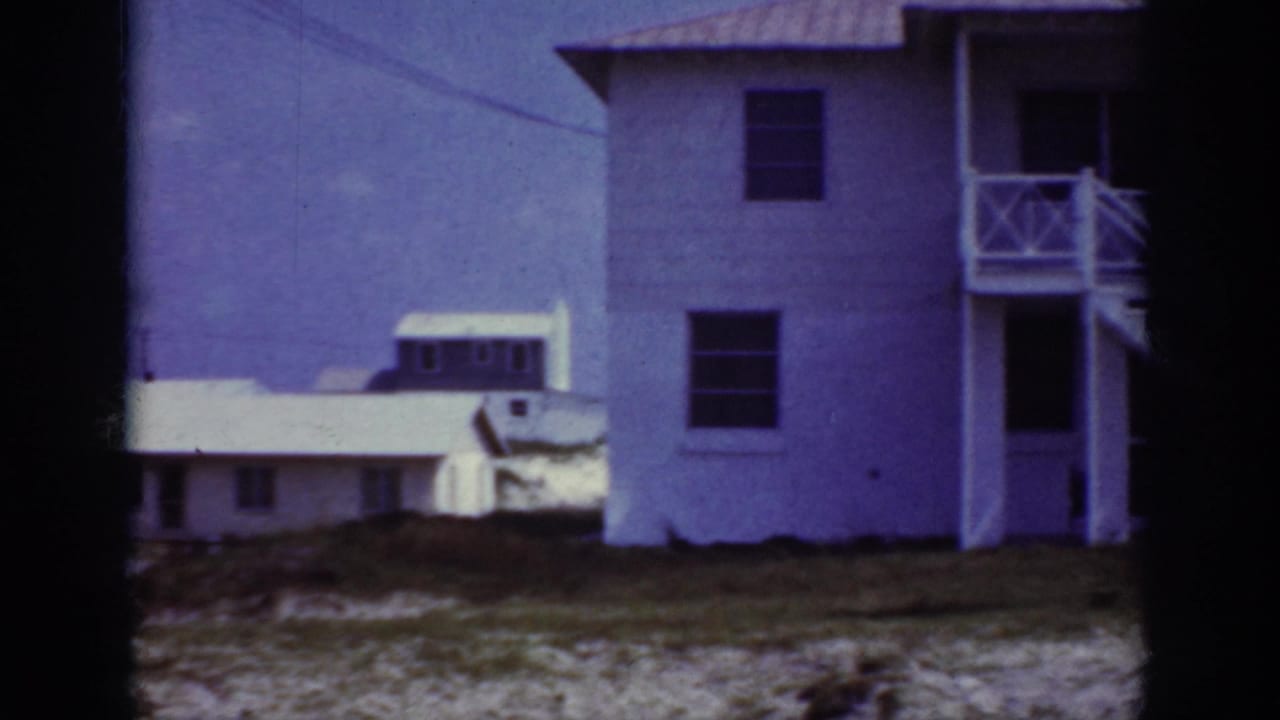 Side view of a white two-story house with balcony railings and another low house behind it.