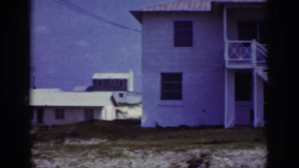 1962 Raleigh frame showing the side of a white two-story house, balcony railings, dark windows, sandy ground, and a smaller residence in the background.