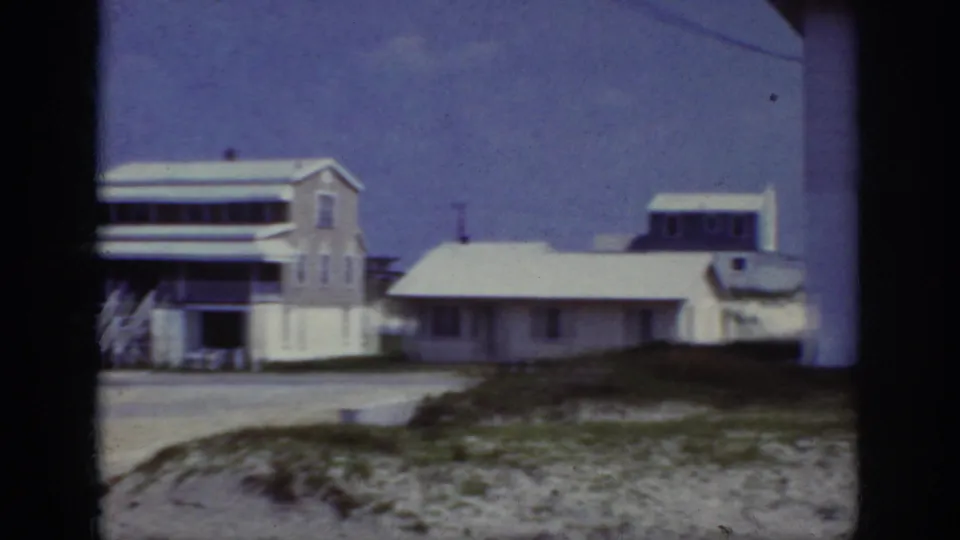 1962 Raleigh home-movie view of low white houses and a taller residence beyond sandy roadside ground, grass patches, and a blue-purple sky.