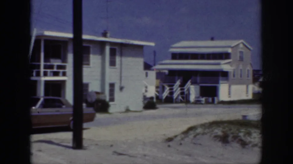 1962 Raleigh frame showing a quiet residential street with white houses, exterior stairs, utility poles, sandy ground, scrub grass, and a parked car.