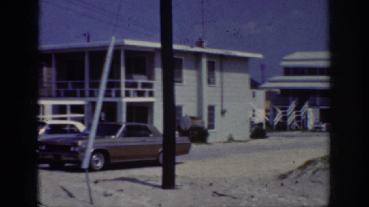 1962 home-movie frame of parked cars outside pale residential buildings in the Raleigh, North Carolina clip metadata context.