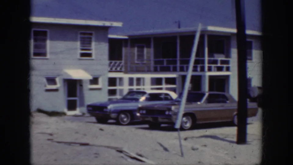 The frame shows Raleigh-context 1962 residential buildings with parked cars, balcony rails, sand, and utility poles.