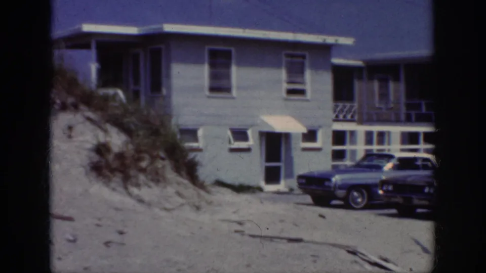 A 1962 Raleigh-labeled home-movie frame shows a pale two-story residence beside sand, dune vegetation, and parked cars.