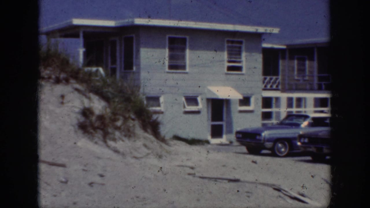 1962 home-movie frame showing a light blue beach house, sandy dune, beach grass, and a parked car in bright sun.
