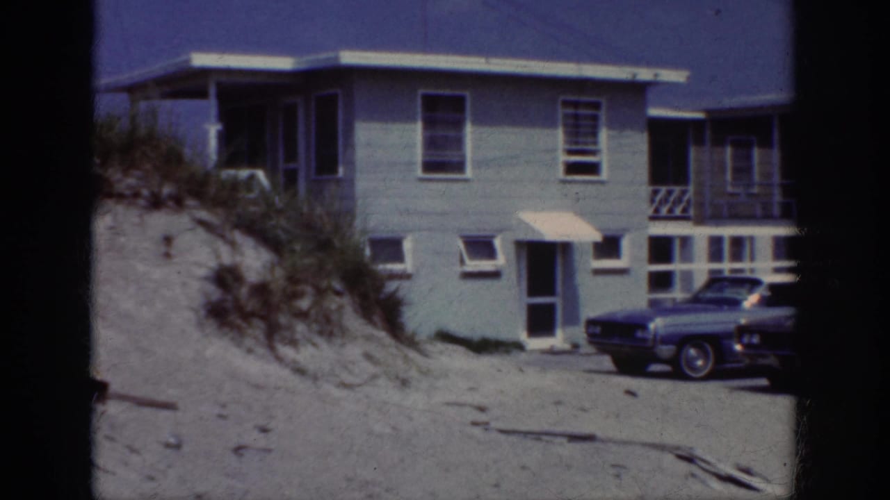 Beach house exterior with sandy foreground and a car parked near the wall.