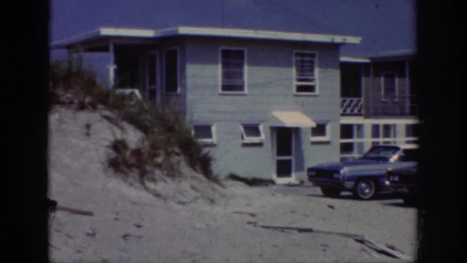 1962 home-movie view of a beach house exterior, sandy dune, small awning, windows, and a parked car.