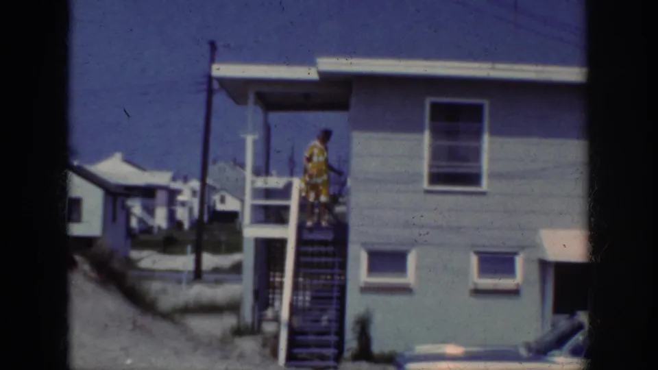 1962 home-movie frame showing a woman in a yellow patterned dress on outside stairs beside a light blue beach house.