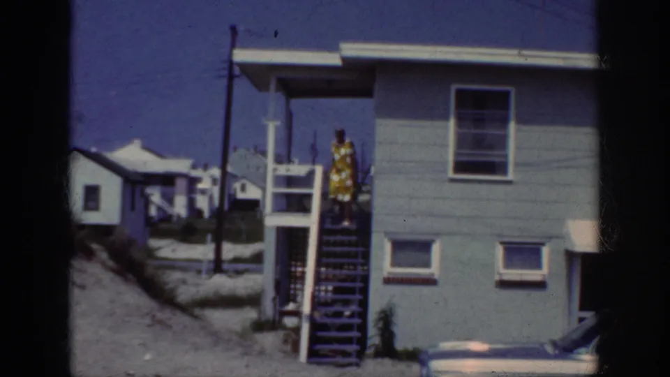 1962 home-movie frame of a woman in a yellow patterned dress walking down outdoor stairs at a coastal house.
