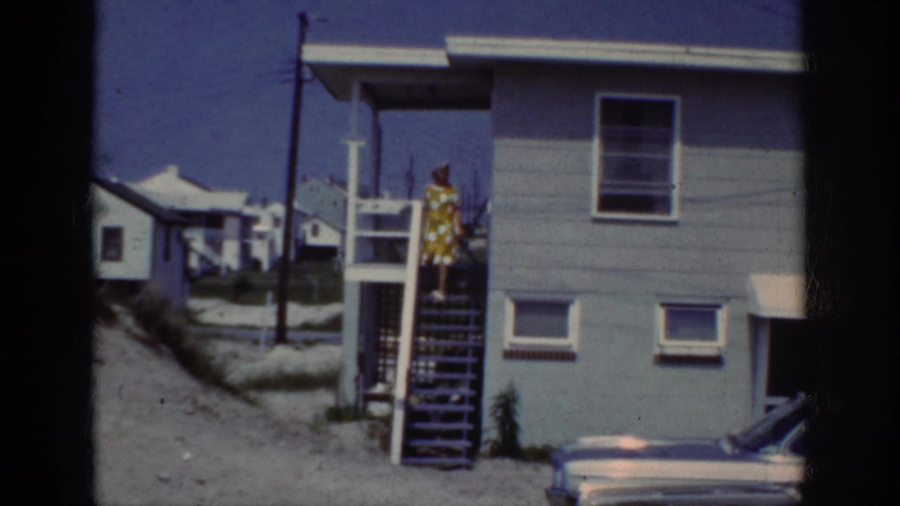 1962 Raleigh home-movie frame showing a woman in a yellow polka dot dress at wooden exterior stairs beside a light blue house.