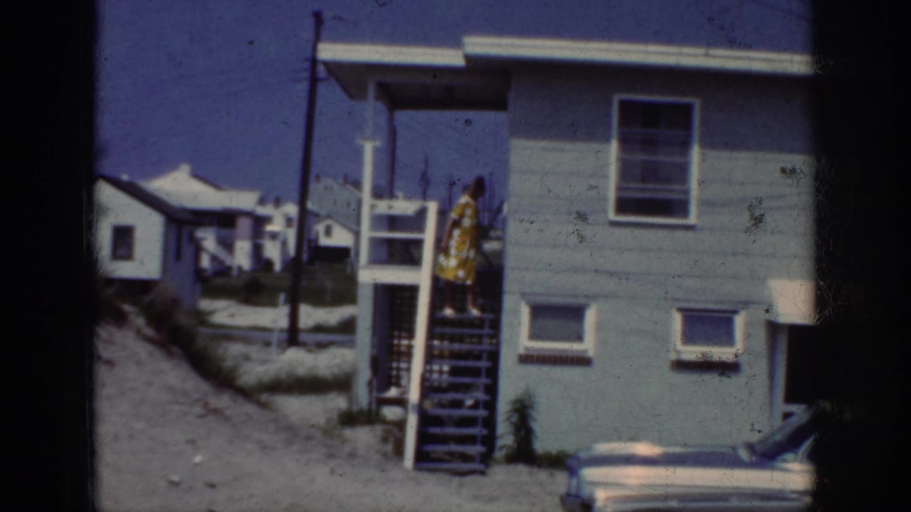 1962 Raleigh frame showing a woman in a yellow polka dot dress walking down exterior wooden stairs near a parked car.