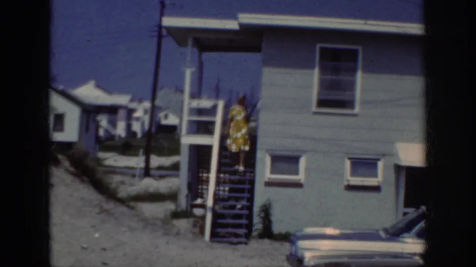 1962 Raleigh home-movie view of a woman in a yellow polka dot dress midway down wooden stairs outside a light blue house.