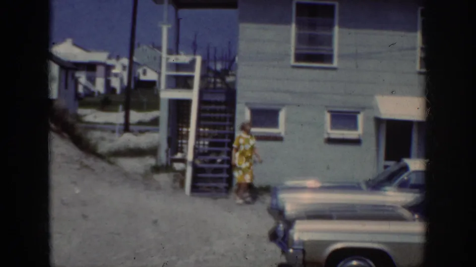1962 Raleigh frame showing the woman reaching the sandy ground below exterior stairs, with a parked car and house behind her.