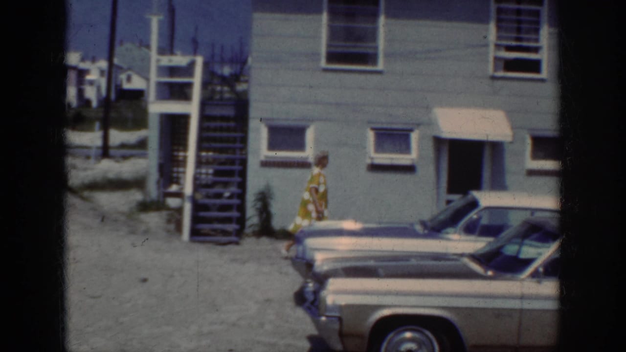 1962 Raleigh home-movie frame showing a woman in a yellow-green patterned dress approaching a house past parked early-1960s cars.