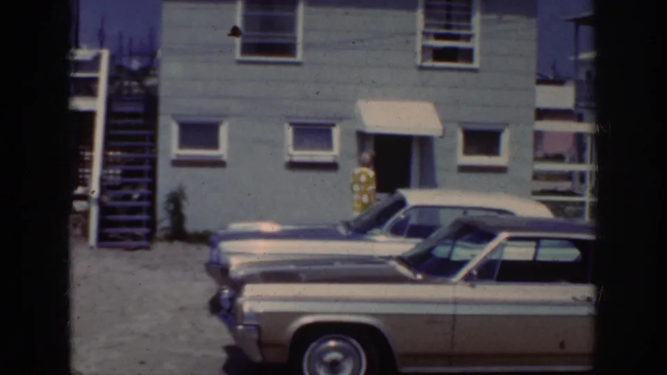 1962 Raleigh exterior frame with a woman at the doorway of a two-story house and early-1960s cars parked in front.