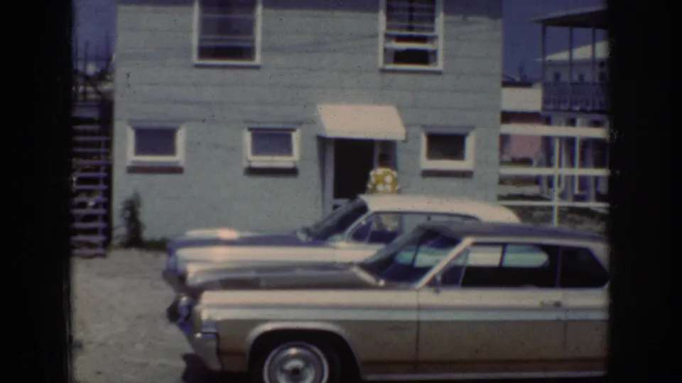 1962 Raleigh home-movie frame showing the woman partly obscured at the doorway, with a station wagon and house exterior visible.