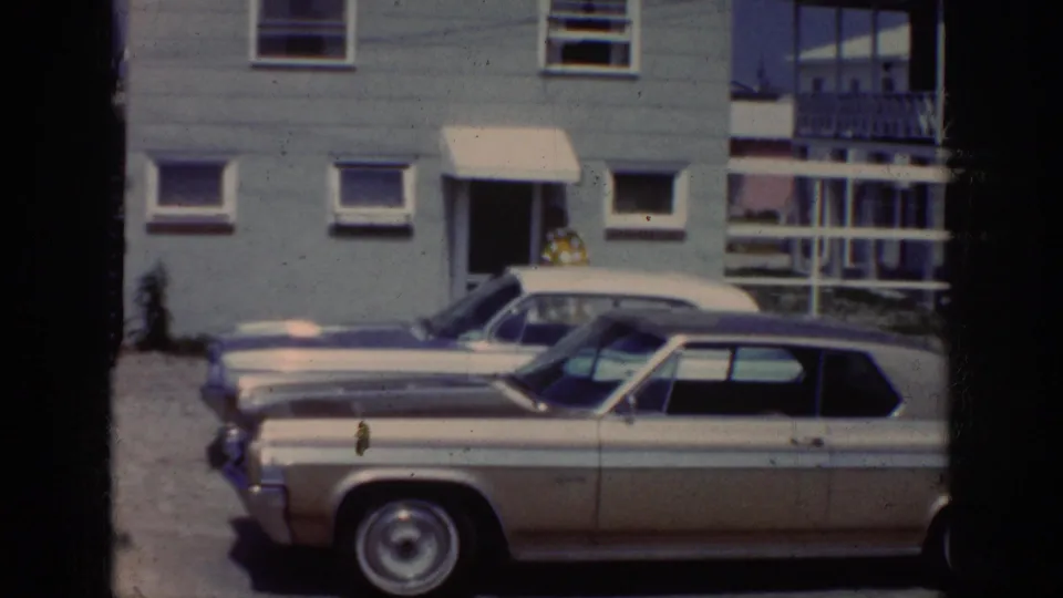 1962 Raleigh exterior frame with early-1960s cars in the foreground and a woman just visible near the house doorway.