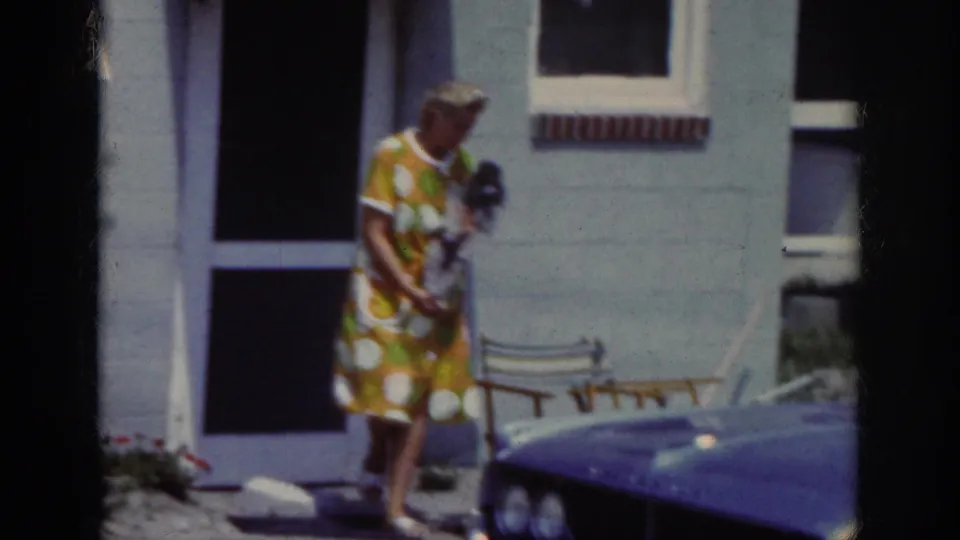 The woman carries the small dog as she shifts position near the screen door and wooden chair in 1962 Raleigh footage.
