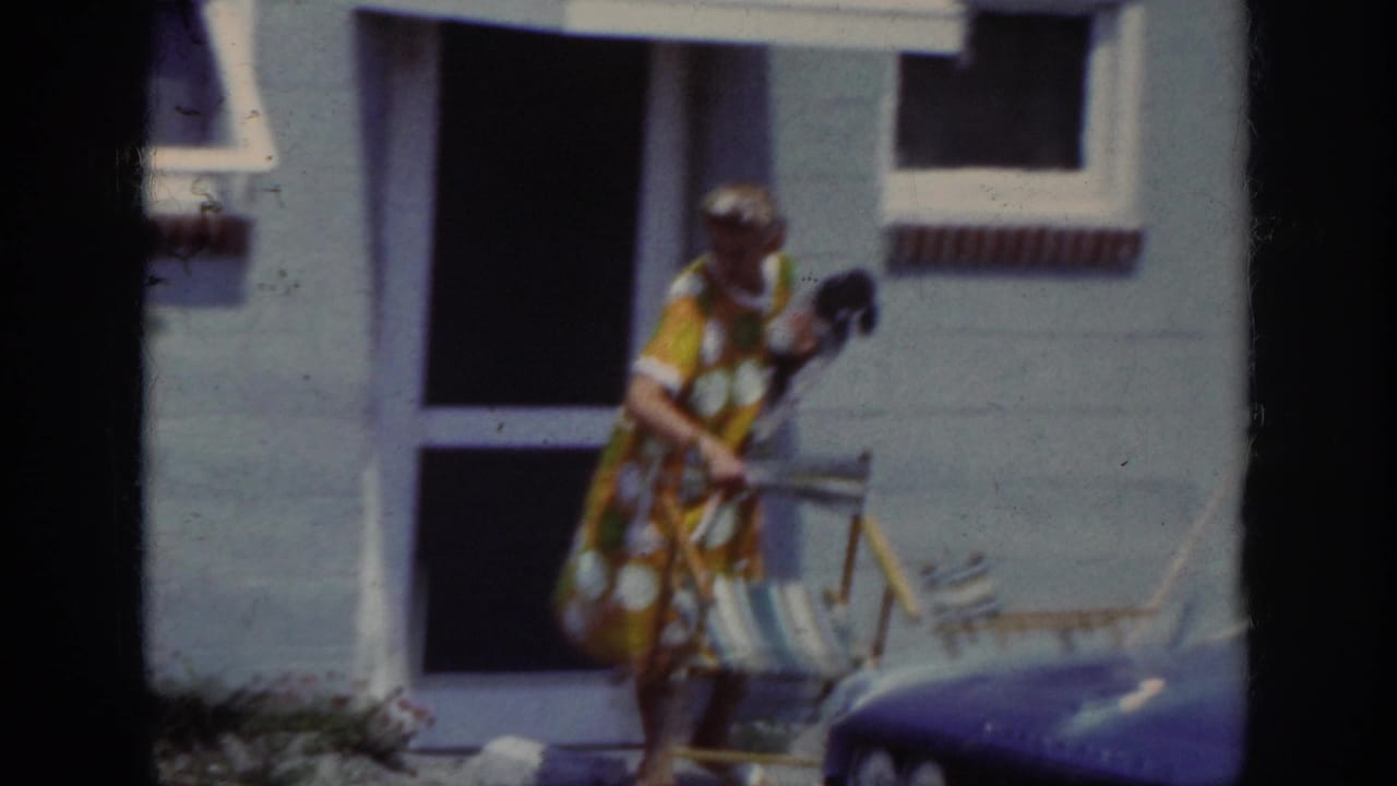 1962 Raleigh home-movie frame of a woman outside a house, holding a small dog while standing by a striped folding chair.