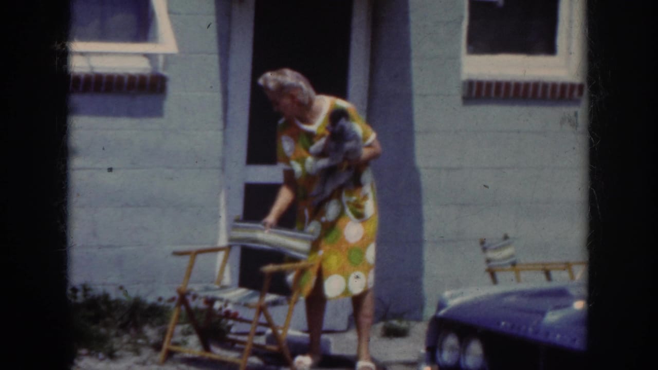 In the Raleigh 1962 home-movie footage, a woman with a small dog arranges a striped folding chair outside a light-colored house.