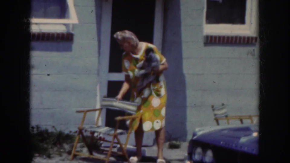 In the Raleigh 1962 home-movie footage, a woman with a small dog arranges a striped folding chair outside a light-colored house.