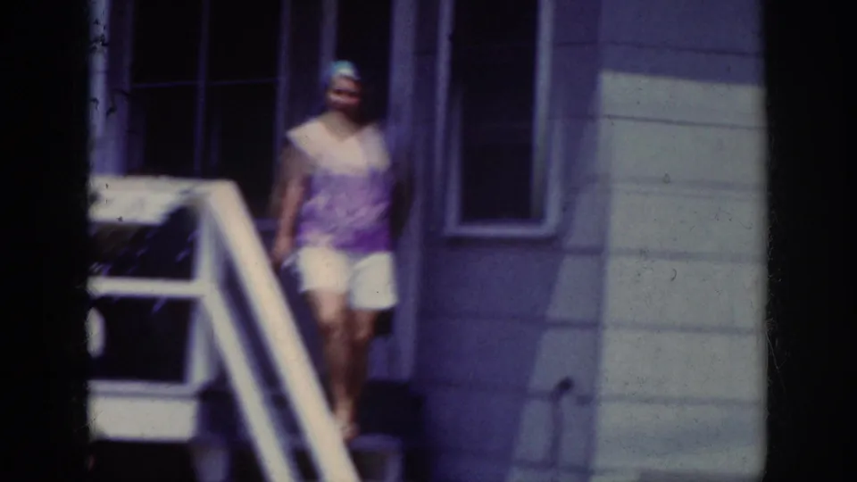 A woman in a purple top and white shorts descends exterior stairs in the 1962 Raleigh home-movie scene.