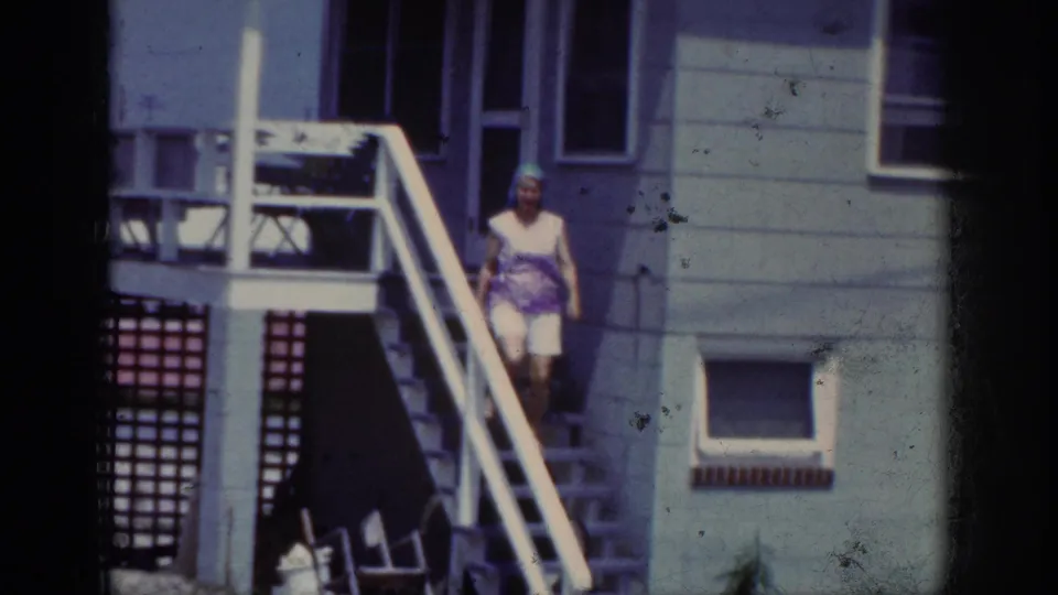 In Raleigh 1962 home-movie footage, a woman in a blue headscarf and white shorts steps down outdoor stairs by a house.