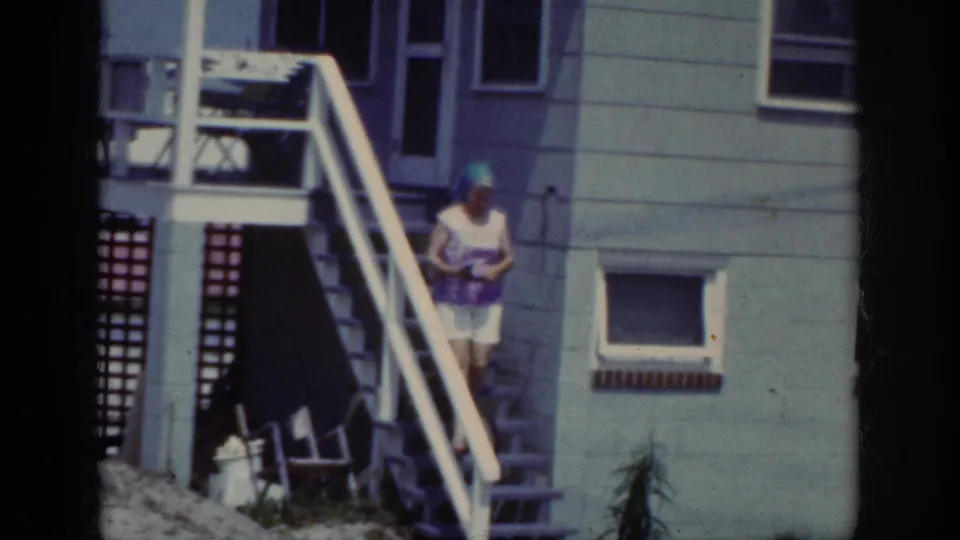 The woman descends the house stairs in Raleigh, wearing a blue headscarf, patterned top, and white shorts.