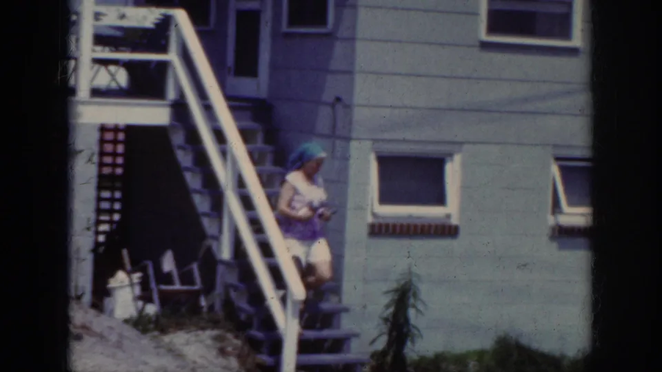 A home-movie frame shows the woman lower on the stairs, carrying a small object beside the house exterior.