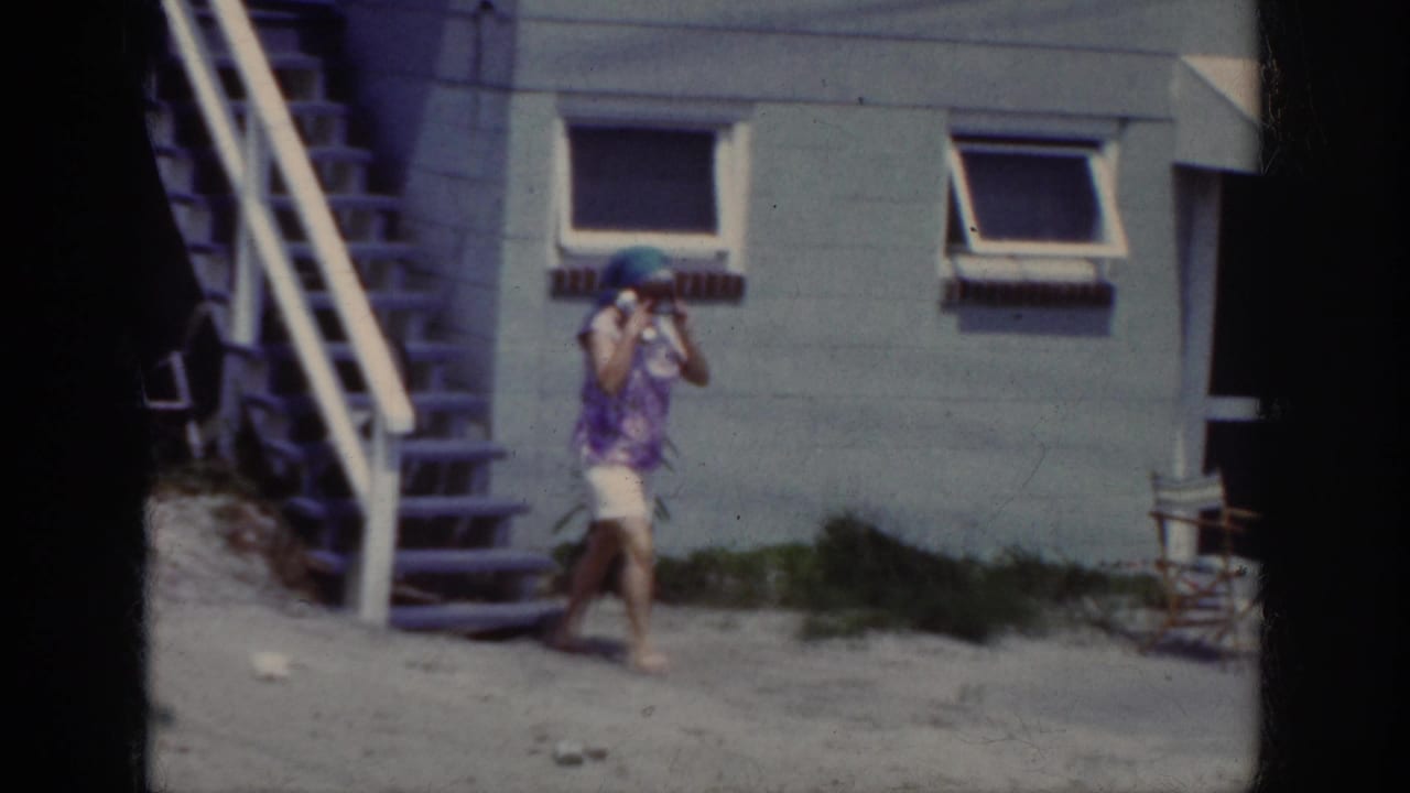 In 1962 Raleigh home-movie footage, a woman in a blue head scarf, sunglasses, purple top, and white shorts walks barefoot near exterior stairs.