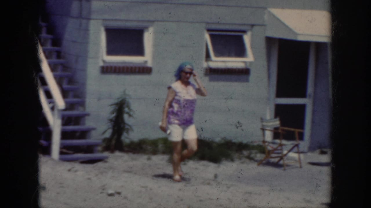 Woman in sunglasses walks across a sandy yard beside a house with open windows and a folding chair.