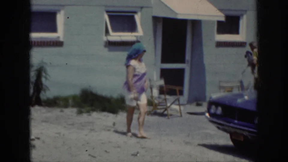 The woman moves past the light blue house and folding chair while the front of a dark period car appears at the right edge.