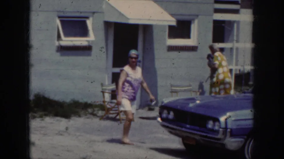 At the end of the clip, the woman walks toward the camera area with a period car in front and another adult figure near the doorway.
