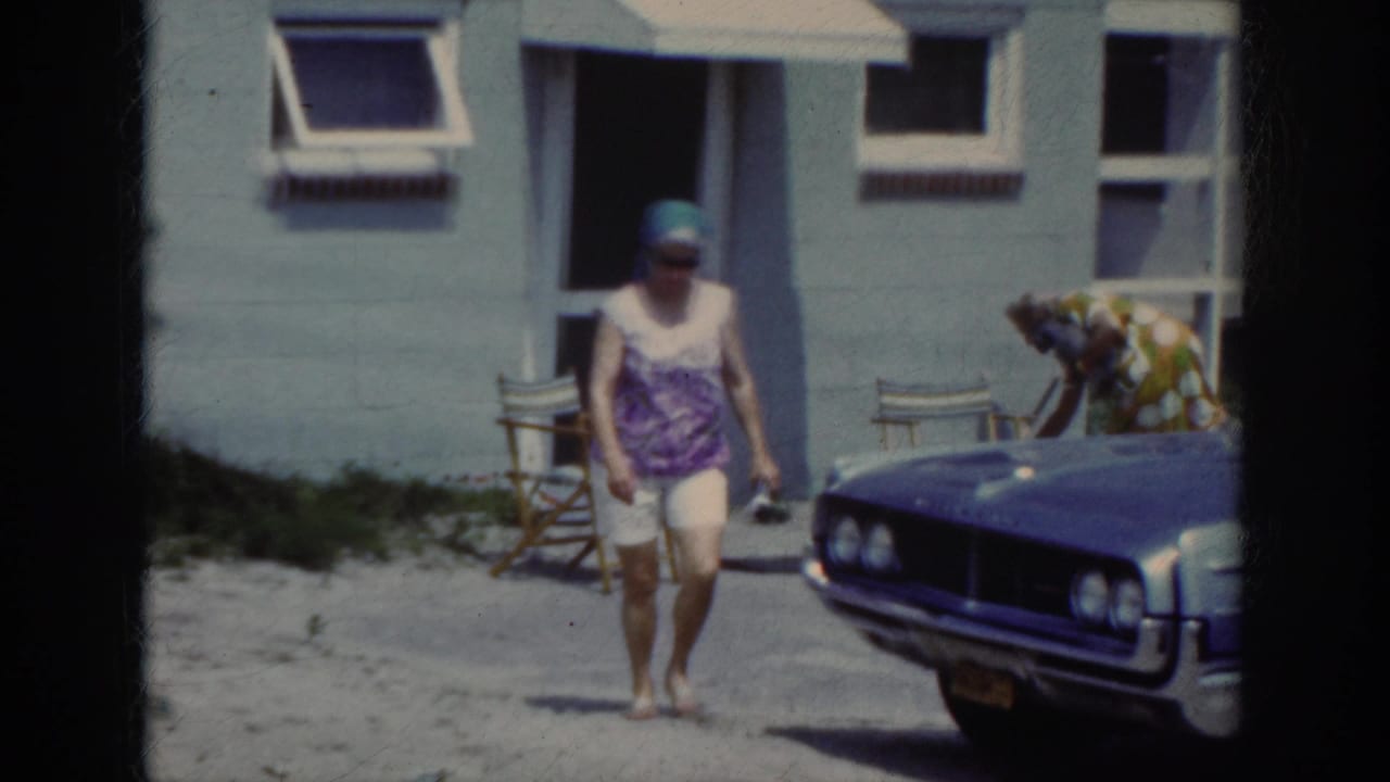 In stated 1962 North Carolina home-movie footage, a woman walks barefoot near a parked blue car and a light-colored building.